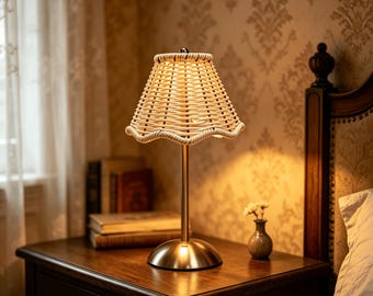 May include: A table lamp featuring a woven light-coloured rattan lampshade with a scalloped edge, supported by a slender metallic gold-toned central rod and a matching circular base, emitting a warm, soft glow that illuminates the surrounding area.