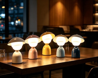 May include: Five mushroom-shaped table lamps arranged in a row, each featuring a wide, rounded shade and a glowing spherical bulb. The lamps are coloured white, light pink, yellow, light blue, and dark blue, each with a smooth, solid base.