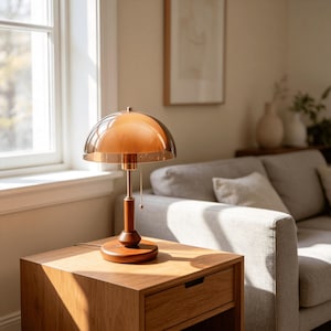 Puede incluir: Una l&aacute;mpara de mesa con una pantalla de cristal &aacute;mbar en forma de c&uacute;pula, un v&aacute;stago de metal fino en tono lat&oacute;n y una base circular de madera. La l&aacute;mpara incluye un peque&ntilde;o interruptor de cadena que cuelga bajo la pantalla.