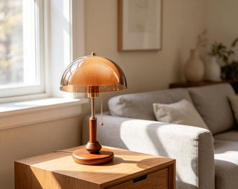 May include: A table lamp featuring a dome-shaped amber glass shade, a slender brass-toned metal stem, and a circular wooden base. The lamp includes a small pull chain switch hanging from the underside of the shade, providing a warm glow.
