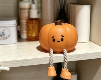 May include: A small orange pumpkin figurine with a brown stem and a sad facial expression. The pumpkin has two dangling legs made of thick, light-coloured twisted rope, each ending in a small orange boot that hangs over the edge of the shelf.