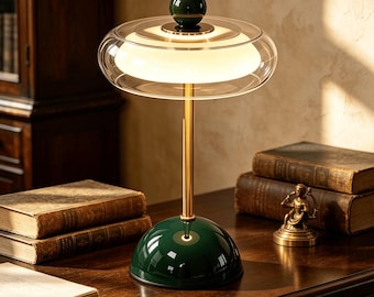 May include: A table lamp featuring a dark green dome base, a slender gold-toned metal stem, and a wide, clear glass shade surrounding a central white glowing light diffuser. A small dark green sphere sits atop the centre of the glass shade.