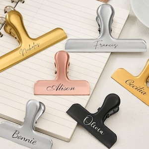 May include: A collection of six metal bulldog clips in gold, silver, rose gold, and black finishes. Each clip is engraved with a name in a cursive script: Nicholas, Francis, Alison, Cecilia, Bonnie, and Olivia. The clips feature a spring-loaded mechanism.