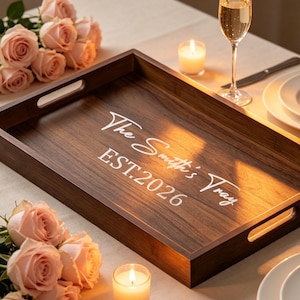 May include: A rectangular dark brown wooden serving tray with cutout handles, featuring the white printed text "The Smith's Tray" and "EST.2026" on the base. The tray is surrounded by light pink roses, two small lit candles, and white plates.