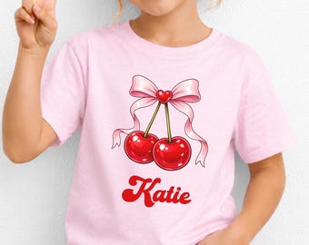 以下が含まれることがあります： 淡いピンク色の半袖Tシャツ。中央に小さな赤いハートが付いた淡いピンクのリボンで結ばれた、鮮やかな赤色のサクランボ2個のグラフィックが描かれています。サクランボの下には、太い赤色のレトロなフォントで「Katie」という名前がプリントされています。