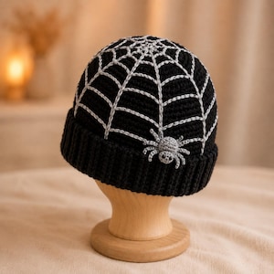 Könnte beinhalten: Eine schwarze Häkelmütze mit einem silbernen Spinnennetz-Muster, das über die Krone gestickt ist. Eine kleine, hellgraue Häkelspinne mit dunklen Augen ist seitlich am Netzdesign befestigt. Die Mütze hat einen dicken, gerippten Saum am unteren Rand.