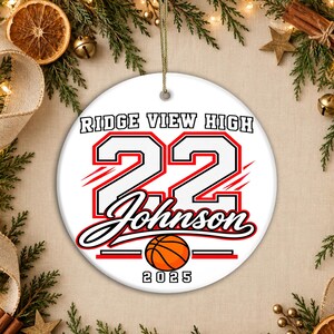May include: A round white ceramic ornament featuring the text Ridge View High, the number 22, the name Johnson, and the year 2025 in black and red lettering. A graphic of an orange basketball is centered below the name, and a gold string is attached.