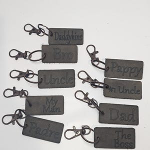 May include: A collection of dark gray leather keychains with various embossed titles, including "Daddykings", "Bro", "Uncle", "My Man", "Padre", "Pappy", "#1 Uncle", "Dad", and "The Boss". Each keychain has a metal clasp.