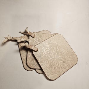May include: A stack of four square, light beige coasters with a detailed, embossed paisley design. Each coaster has a small, attached wooden bird. The coasters are made of a light-colored material.