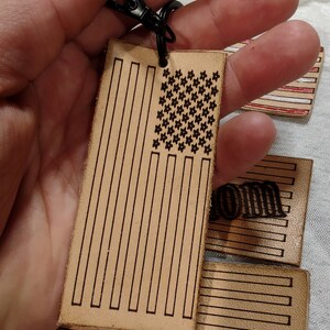 May include: A leather keychain with a black metal key ring and a brown leather tag featuring an American flag design. The flag has red and white stripes and a field of blue stars.