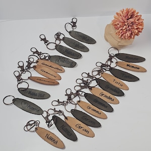 May include: A collection of 17 leather keychains with various names and titles engraved on them. The keychains are oval-shaped and come in two colors: brown and gray. The keychains are arranged in a row on a white surface.