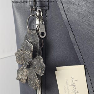 Puede incluir: Un bolso tote de cuero negro con cierre de cremallera y un tirador de cremallera de metal plateado. Un llavero de metal plateado con un charm de flor de cuero decorativo está unido al tirador de la cremallera. El bolso tiene una etiqueta que dice "whispers+touch" Artículos de cuero hechos a mano.