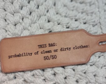 May include: A brown leather tag with the text "THIS BAG: probability of clean or dirty clothes: 50/50" printed on it.