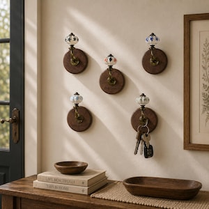 Peut inclure: Cinq patères murales composées de bases rondes en bois foncé et de ferrures en laiton surmontées de boutons en céramique décoratifs. Chaque bouton présente un motif floral unique en bleu, noir ou orange. Un crochet porte un trousseau de clés.