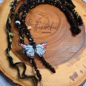 May include: Black dreadlock extensions with decorative beads, green accents, and a blue butterfly charm. The extensions are arranged on a wooden surface, showcasing the artisan jewelry design.