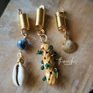 May include: Three gold-toned hair accessories with unique charms. One features a blue and white speckled bead and a white seashell. Another has a braided beige element with green beads. The third has a gold seashell charm. The accessories are artisan jewelry.