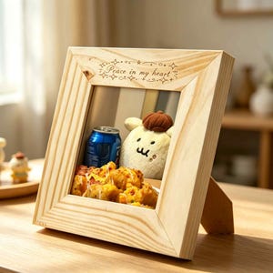 May include: A square light-colored wooden frame with a stand, featuring the engraved text "Peace in my heart" at the top. Inside the frame, a blue aluminum soda can sits next to a small, cream-colored plush toy and a pile of golden-brown food.