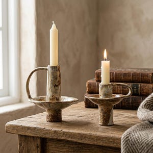 Peut inclure: Deux bougeoirs en céramique avec un vernis moucheté brun et crème, chacun doté d'une poignée latérale et d'une large base circulaire. L'un contient une bougie effilée crème éteinte, l'autre une bougie effilée crème allumée.