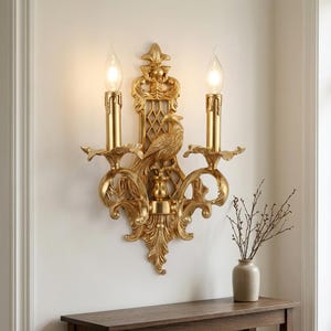 May include: A gold-colored wall sconce featuring two candle-style light fixtures, a central bird figure, and an intricate lattice backplate with ornate scrollwork. The fixture is mounted on a wall and emits a warm glow from two bulbs.