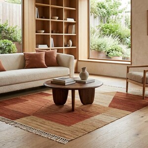 Op de afbeelding: Een rechthoekig vloerkleed met een geometrisch patroon in tinten terracotta, roest en beige, afgewerkt met witte franjes aan de korte zijden. Het kleed ligt op een lichte houten vloer in een kamer met een bank, fauteuil en salontafel.