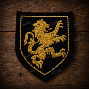 Op de afbeelding: Een zwart schildvormig embleem met een geborduurde gouden leeuw in een klimmende houding. Het embleem is omlijnd met een dikke, gedraaide gouden koordrand. Het woord MILITOX is herhaald als een vaag watermerk over de afbeelding heen.