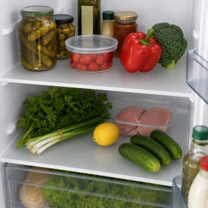 Può includere: L'interno di un frigorifero con due ripiani contenenti vari alimenti. Il ripiano superiore contiene barattoli di cetriolini e olive, un contenitore di pomodorini, un peperone rosso e broccoli. Il ripiano inferiore contiene erbe fresche, cipollotti, un limone, carne cruda in un contenitore trasparente e cetrioli.