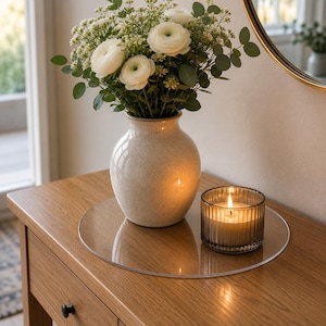 Può includere: Un vassoio circolare trasparente in acrilico che sostiene un vaso in ceramica color crema maculato, riempito con fiori di ranuncolo bianchi e foglie di eucalipto verdi, accanto a un portacandele in vetro scanalato con una candela bianca accesa.
