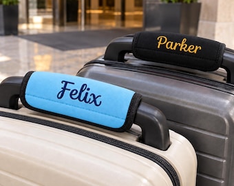 May include: Two luggage handle wraps attached to suitcase handles. One wrap is light blue with the name Felix embroidered in dark blue script. The other wrap is black with the name Parker embroidered in yellow script. Both wraps have black trim.