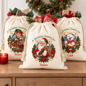 Pode incluir: Três sacos de presente de lona na cor creme com cordões grossos. Cada saco apresenta uma ilustração festiva dentro de uma moldura decorativa, o texto A Christmas Surprise for e um nome: Mason com um elfo, Nolan com o Pai Natal e Caleb com um urso.