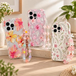 May include: Three smartphone cases with attached beaded wrist straps. The left case is clear with large pink and yellow hibiscus flowers. The center case is clear with pink hibiscus flowers and seashells. The right case is white with small pink flowers.
