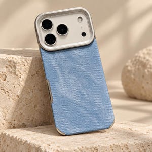 May include: A smartphone case featuring a light blue, textured fabric back panel with a diagonal weave pattern. The upper section includes a metallic silver frame surrounding the camera module, which houses three circular lenses and a flash.