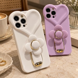 May include: Two smartphone cases, one white and one lavender, each featuring a heart-shaped camera cutout and a flower-shaped ring holder with gold-tone accents. The cases have a smooth, glossy finish and a raised heart design around the camera lens.