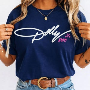 May include: A navy blue short-sleeved t-shirt featuring the white cursive text Dolly across the chest, accompanied by the pink handwritten text xoxo and three small pink hearts. The shirt is worn with a brown leather belt and blue denim jeans.