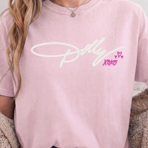 May include: A light pink short-sleeve t-shirt featuring the white cursive text Dolly across the chest. Below the text, the word XOXO is printed in bright pink, accompanied by three small pink hand-drawn hearts of varying sizes.