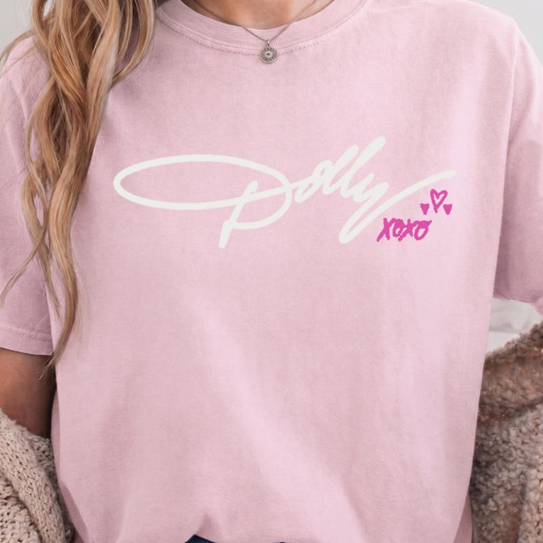 May include: A light pink short-sleeve t-shirt featuring the white cursive text Dolly across the chest. Below the text, the word XOXO is printed in bright pink, accompanied by three small pink hand-drawn hearts of varying sizes.