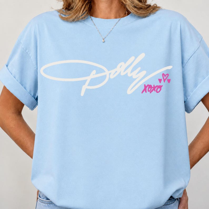 May include: A light blue short-sleeve t-shirt featuring the white cursive text Dolly across the chest. Below the text, the word xoxo is printed in pink, accompanied by three small pink heart icons. The shirt has a crew neckline and rolled sleeves.