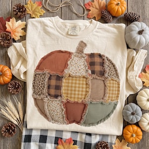Könnte beinhalten: Ein cremefarbenes Kurzarm-T-Shirt mit einer großen, zentralen Kürbisgrafik, die aus verschiedenen Stoffflicken in Orange-, Braun-, Beige- und Salbeigrüntönen besteht, mit einem Comfort Colors-Markenetikett am inneren Halsausschnitt.