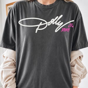 May include: A charcoal gray short-sleeve t-shirt featuring the white cursive text Dolly across the chest. Below the text, the word xoxo is printed in bright pink, accompanied by two small pink hearts. The shirt has a crew neckline and a relaxed fit.