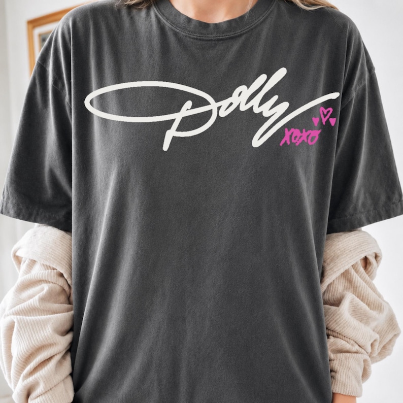 May include: A charcoal gray short-sleeve t-shirt featuring the white cursive text Dolly across the chest. Below the text, the word xoxo is printed in bright pink, accompanied by two small pink hearts. The shirt has a crew neckline and a relaxed fit.
