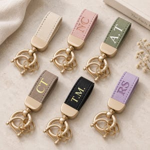May include: Six keychains with textured straps in cream, pink, olive green, taupe, black, and lavender. Each strap features gold-tone hardware and a metal ring. The straps are embossed with the initials NC, A.T, CT, T.M., and RS in various colours.