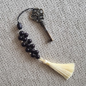 May include: A decorative accessory featuring an antique bronze-colored metal key with an ornate bow, attached to a black cord with a cluster of dark brown spherical beads and small silver-toned spacers, ending in a cream-colored fabric tassel.