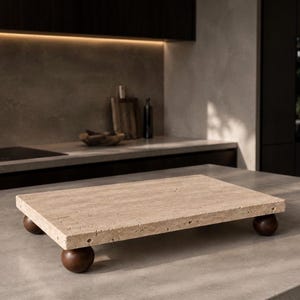 Pode incluir: Uma bandeja retangular de pedra travertino bege com uma superfície porosa e texturizada, apoiada sobre quatro pés esféricos de madeira castanho-escura. O suporte oferece uma plataforma elevada que destaca os padrões naturais da pedra.