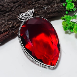Könnte beinhalten: Ein großer, birnenförmiger Anhänger mit einem facettierten, tiefroten Edelstein in einer dekorativen Fassung aus Sterlingsilber. Die Silbereinfassung weist an den Rändern filigrane Metallarbeiten und eine detaillierte, verzierte Öse auf.