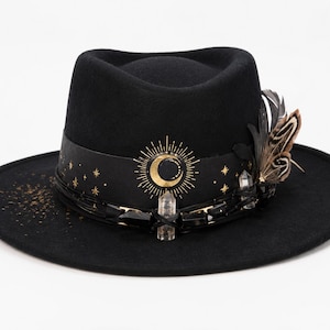 May include: A black fedora hat featuring a wide brim and a black hatband decorated with a gold sun and crescent moon graphic, small stars, clear quartz crystals, black beads, and a cluster of brown and black patterned feathers on the side.