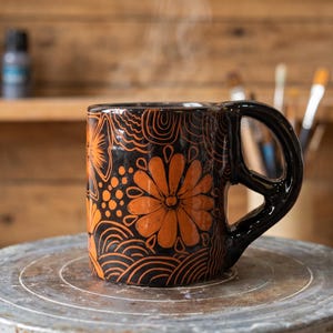 May include: A black ceramic mug featuring a hand-carved orange floral and wave pattern. The mug has a large, thick, rounded handle and is emitting a thin wisp of steam from the top, indicating a hot beverage inside the vessel.