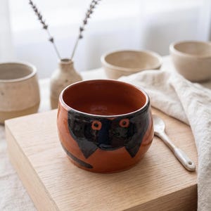 May include: A round terracotta-colored ceramic bowl featuring a hand-painted black cat face design with two orange circular eyes. The bowl has a smooth, glossy finish and a wide opening, showcasing a deep interior and a simple, rounded silhouette.