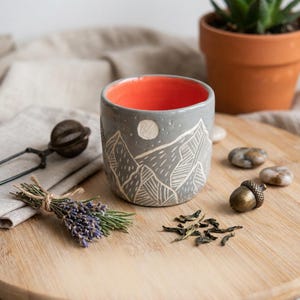 May include: A ceramic cup with a gray exterior featuring a white etched mountain and moon design, and a bright orange interior. The cup is displayed alongside a metal tea infuser, a bundle of dried lavender, and a small metal acorn ornament.