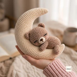 Pode incluir: Um pequeno brinquedo de crochê feito à mão representando uma lua crescente bege clara com um urso castanho a dormir sobre ela. O urso tem olhos fechados bordados, um nariz castanho escuro e orelhas pequenas, com acabamento em fio texturizado.