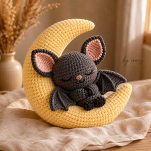 May include: A dark gray crocheted bat with pink inner ears and rosy cheeks, sleeping while nestled in the curve of a large, textured, pale yellow crocheted crescent moon. The bat has small wings and is positioned in a curled, resting posture.