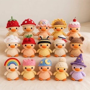Puede incluir: Una colección de quince patitos de ganchillo dispuestos en tres filas. Cada patito presenta un sombrero o accesorio de ganchillo único, que incluye una fresa, una seta, un girasol, una cereza, una abeja, un arcoíris, una corona y un sombrero de mago.